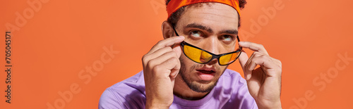 annoyed african american man in eyeglasses and headband rolling eyes on orange background, banner photo