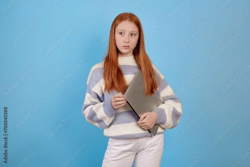 Confident redhead teenage girl holding laptop on blue background in studio. Confident redhead teenage girl holding laptop on blue background in studio.