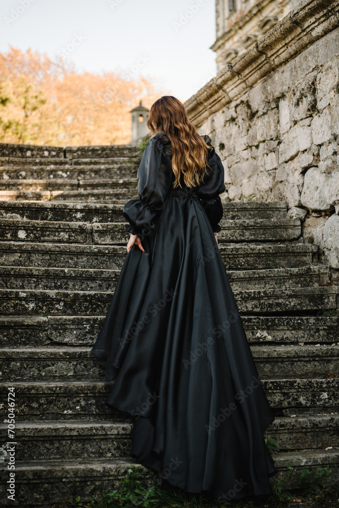 Female in long black dress walk on stairs near ancient palace at sunset. Model girl goes in nature autumn day. Luxury woman near old Pidhirtsi Castle, Lviv region, Ukraine. Stylish bride. Back view