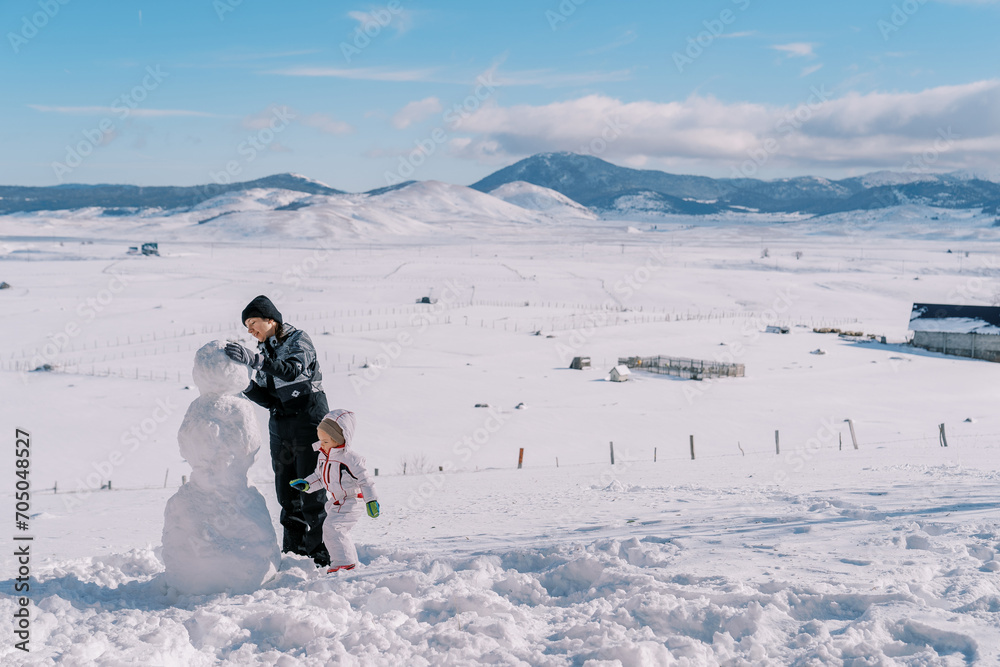 Little girl and her mother are making a snowman in a village in a mountain valley. Side view