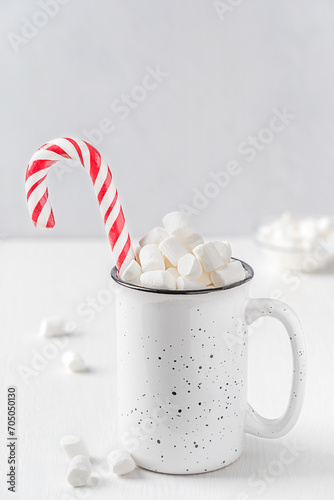 Homemade sweet hot chocolate or traditional cocoa drink decorated with marshmallow topping and striped candy cane served in ceramic mug or cup on white wooden table for cozy winter holiday breakfast