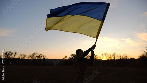 Male ukrainian army soldier going and waving flag of Ukraine on meadow at dusk. Young man in camouflage uniform walking with raised blue-yellow banner as symbol of victory against russian aggression
