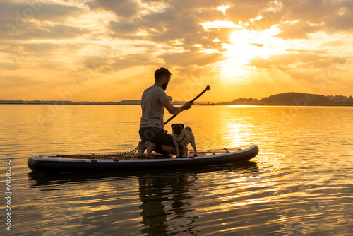 Man paddle boarding at lake during sunset together with pug dog. Concept of active tourism and supping with pets. Brave Dog Standing on SUP Board and enjoy lifestyle on summer petfriendly vacation