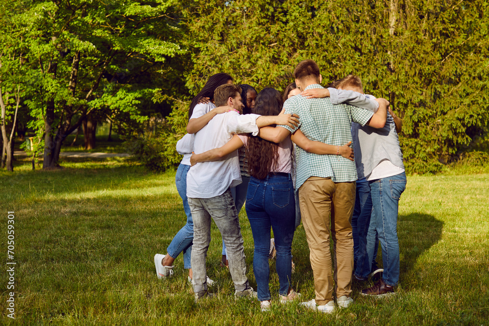 Fototapeta premium Team of young diverse people huddling in park all together. Group of friends standing and hugging on fresh green lawn in beautiful summer park. Teamwork, support, friendship concept