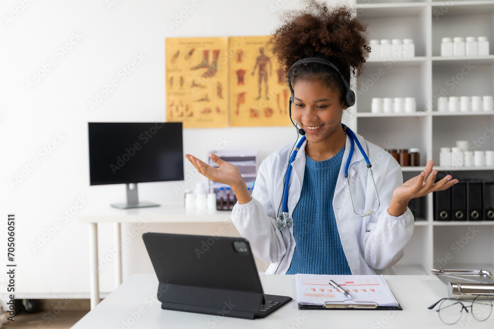 A young African American female doctor in a white coat is sitting at a consulting table using a laptop. Telemedicine service Doctor chats with patient via online video conference