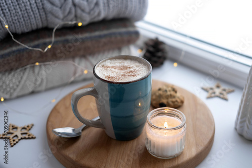 Winter windowsill still life. Blue ceramic cup of hot coffee on window sill. Christmas decorations on the background. Cozy home picture. Warm woolen knitted sweaters, Burn Candle, Cookies. Stock photo