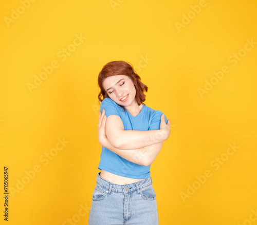 Hugging herself, close up portrait of young attractive cheerful woman enjoying hugging herself. Lovely charming red bob haired girl wear blue t-shirt, jeans. Isolated yellow background.