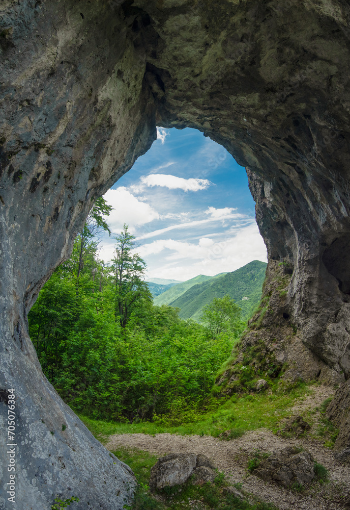 Obraz premium Viewpoint above a deciduous forest and mountains through a cave, eroded in a calcareous cliff on a mountain side. The tunnel has its stone walls covered with moss and lichen. Natural phenomenon.