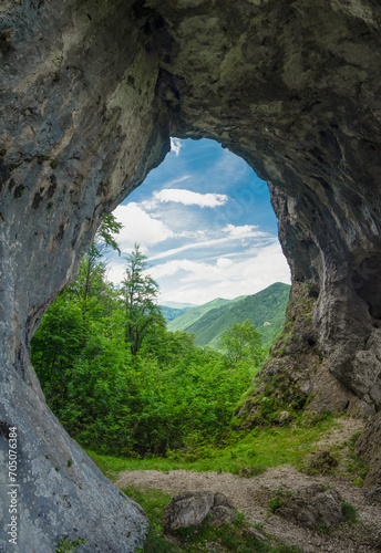 Viewpoint above a deciduous forest and mountains through a cave, eroded in a calcareous cliff on a mountain side. The tunnel has its stone walls covered with moss and lichen. Natural phenomenon.