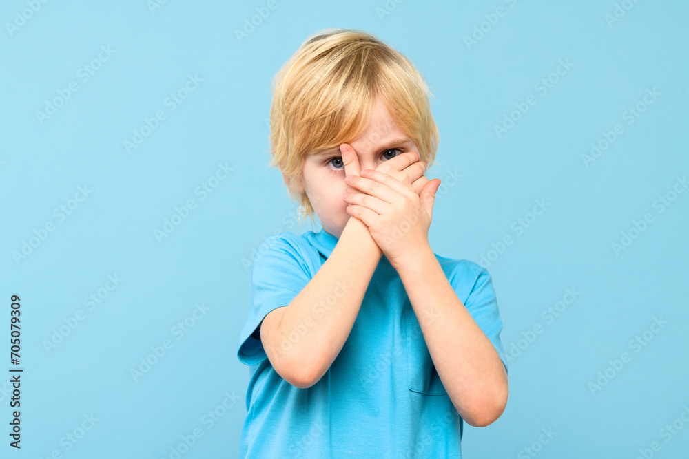 Emotional young boy covering face with hands isolated over pastel blue background. Mental wellbeing and safety in childhood age. Emotional young boy covering face with hands isolated over pastel blue background. Mental wellbeing and safety in childhood age.