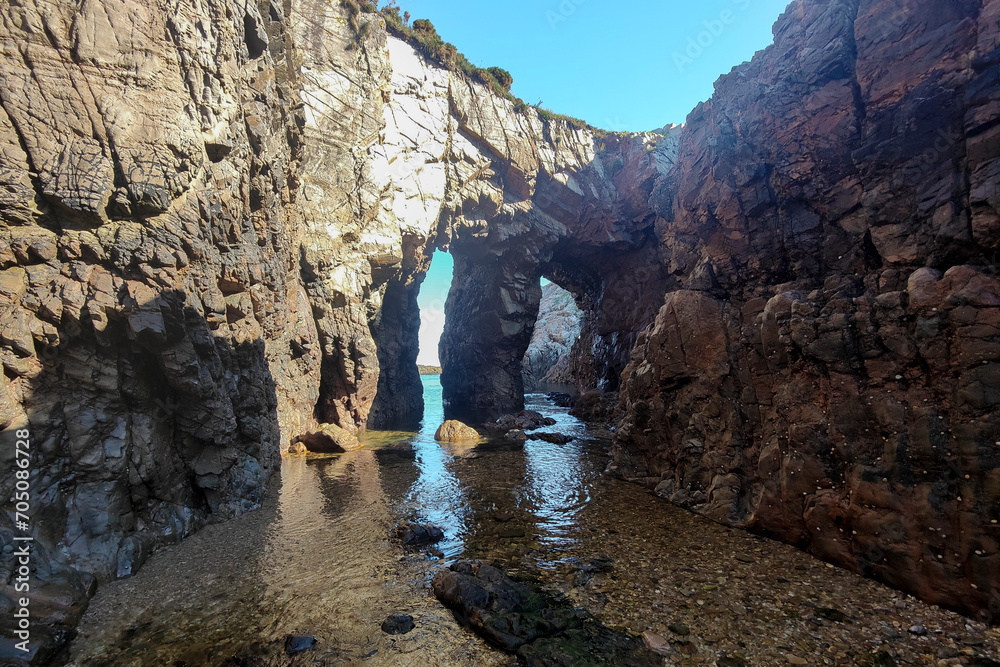 a rocky coastline with natural arches, calm waters reflecting sunlight, under a clear blue sky in asturias