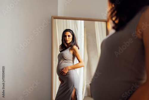 Close up of female portrait of beautiful pregnant in grey dress touching her belly. Reflection of young woman expecting baby in large rectangular mirror. photo