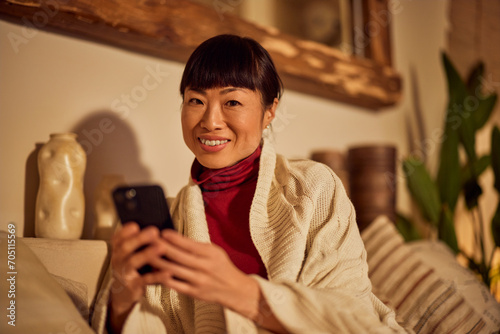 Portrait of a smiling Asian woman, using a mobile phone, looking at the camera while resting on the couch.