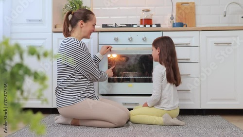 Brown haired mother and daughter sitting on floor near oven waiting for tasty dessert together talking and laughing enjoying family moments on weekend photo