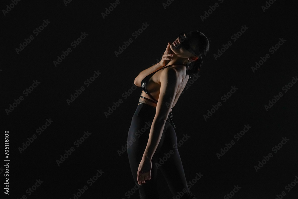 Young woman in black bodice and tights poses on black backlit studio background. Female dancer demonstrates elements of dance on high heels. Dance promo video concept.