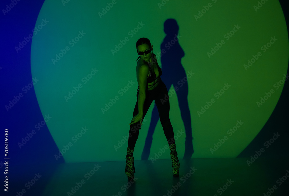 Young woman poses in a dark studio with blue green light as the center of spotlight. The shadow of her body is reflected on the wall. A female dancer demonstrates elements of dance in high heels.
