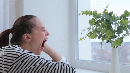 Young Caucasian woman yawning while sitting at window looking outside with bored facial expression being lonely and sad has boring weekend lone at home photo