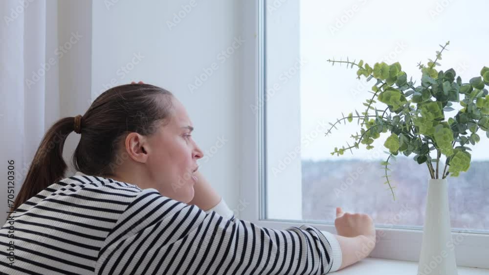 Sad bored woman with brown hair sitting near window touching glass yawning being sleepy tired and lonely female wearing striped shirt spending boring time at home Sad bored woman with brown hair sitting near window touching glass yawning being sleepy tired and lonely female wearing striped shirt spending boring time at home