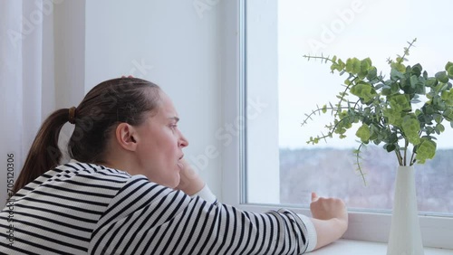 Sad bored woman with brown hair sitting near window touching glass yawning being sleepy tired and lonely female wearing striped shirt spending boring time at home photo