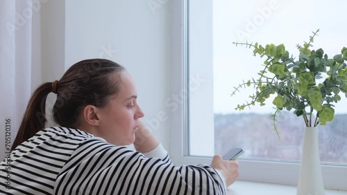 Bored sad woman wearing striped shier sitting near window with her smartphone using mobile app checking emails and social networks trying to entertain herself with internet photo