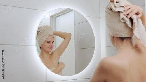 Female wellness. Body treatment. Wellbeing routine. Caucasian woman wrapped in towel on her head looking at mirror at her reflection saving her armpit with razor standing in bathroom photo