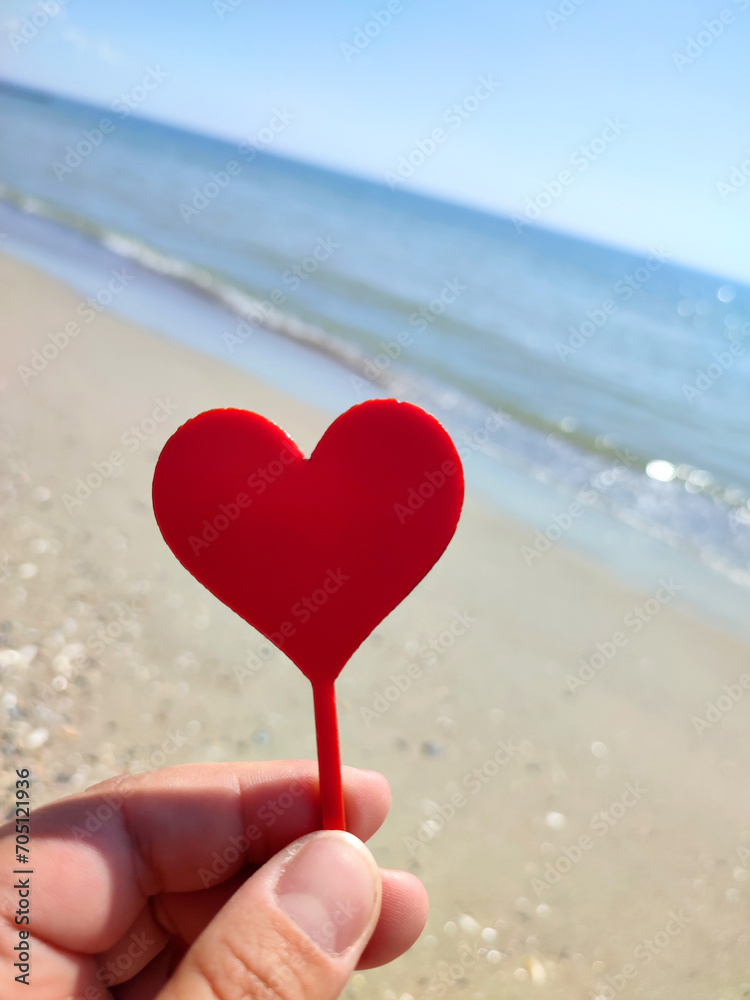 Fototapeta premium Person holding in fingers hand stick in shape red heart on background sea and sea waves, blue sky in seashore on sunny summer day close-up. Concept love romance amour St Valentines Day