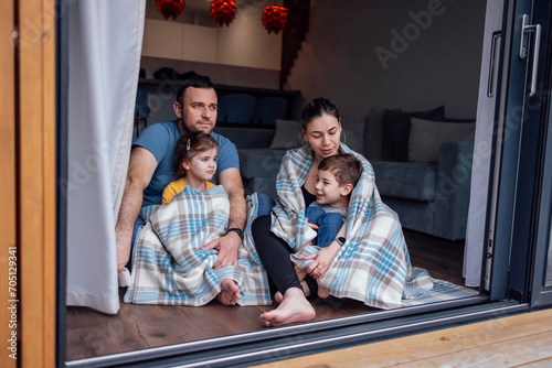 Young parents and small children sit on floor of living room at home in front of open panoramic window.