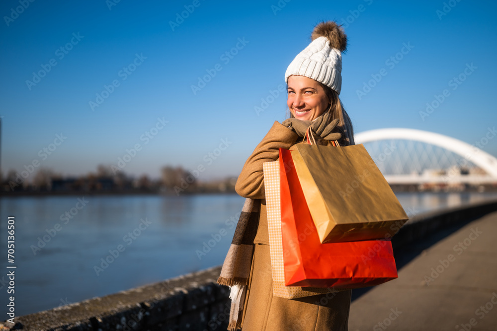 Happy woman in warm clothing with shopping bags enjoys standing by river on sunny winter day.	