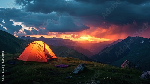Glowing orange tent in the mountains under dramatic evening sky. Red sunset and mountains in the background. Summer landscape