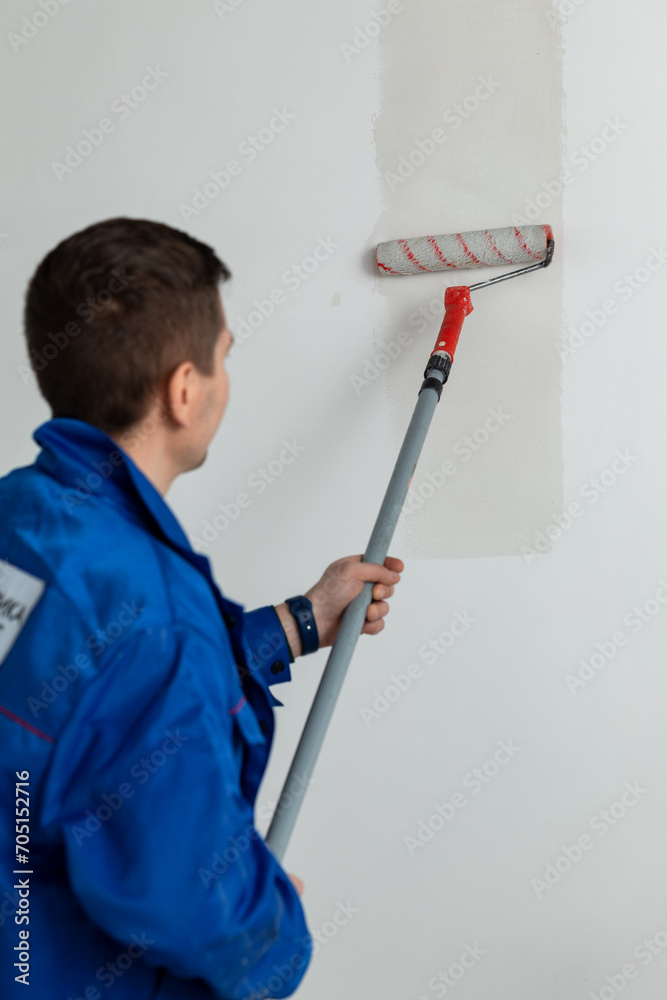 A worker in a blue work jacket applies glue to the wall with a roller for wallpapering A worker in a blue work jacket applies glue to the wall with a roller for wallpapering