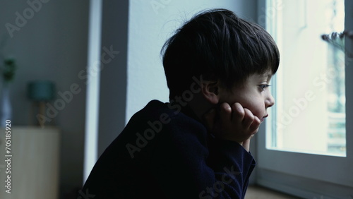 Thoughtful bored child standing at home with hand in chin observing view from apartment window with introspective pensive expression