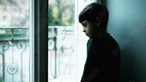 One young boy struggles with mental illness at home with green tint color. Child depressed standing by window with blank expression depicting a meaningless life photo