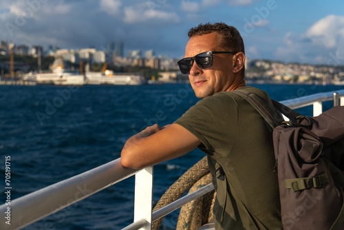 Happy handsome young male tourist in sunglasses with a backpack on his shoulders enjoys the sea and the city from a ferry in Istanbul on a sunny day. Concept of travel, tourism