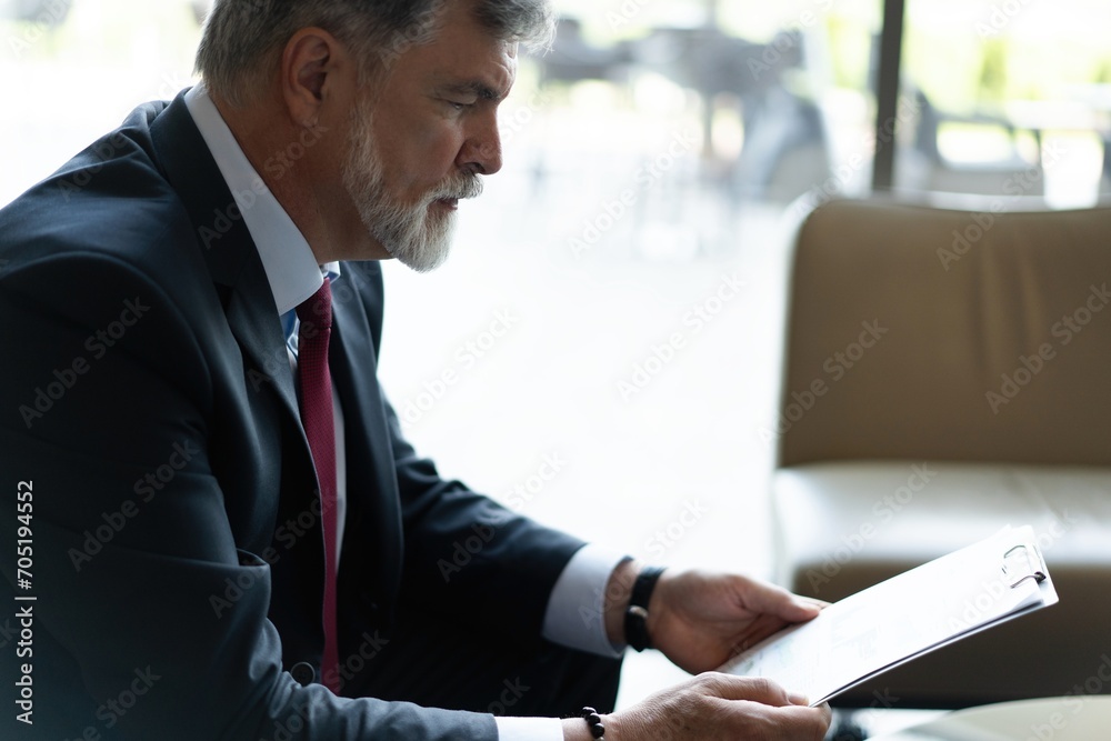 Mature businessman in formal suit concentrating on reading the report, sitting in the lobby and preparing for the meeting.