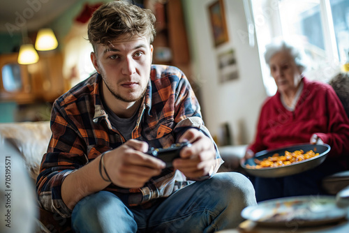 NEET young man not studying and unemployed playing video games, grandmother blurred in background with food photo