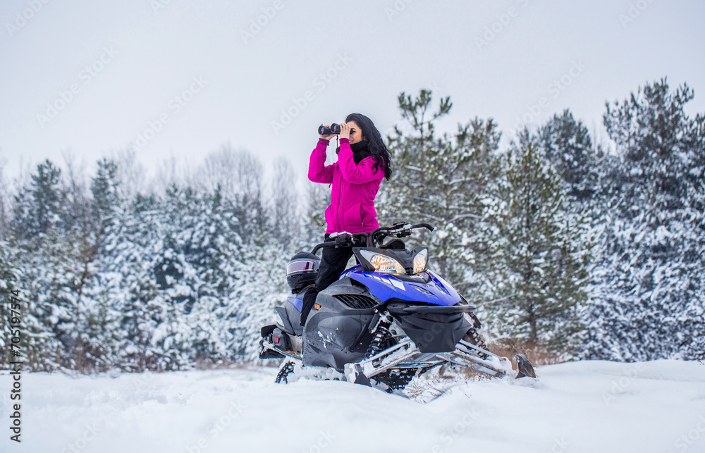 Girl on sports snowmobile in forest. Rides a snowmobile in the mountains. Snowmobile in snow. Woman is riding snowmobile. Travel, girl looks through binoculars. Woman in winter forest with binoculars