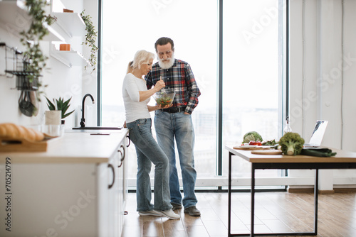 Devoted elderly woman feeds man with gray beard with fresh healthy salad in glass bowl. Happy old pensioner couple having breakfast in modern light kitchen with panoramic windows.