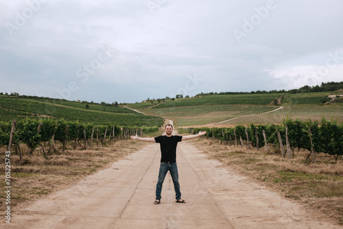 a happy young man dressed in a black T-shirt stands between two fields with grapes. His arms are spread to the side. Pride in the harvest. photo