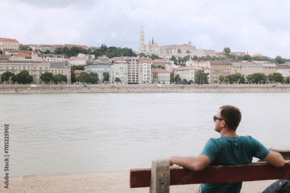 A young man sits on a bench on the Danube River embankment and looks at the panorama of the city of Budapest