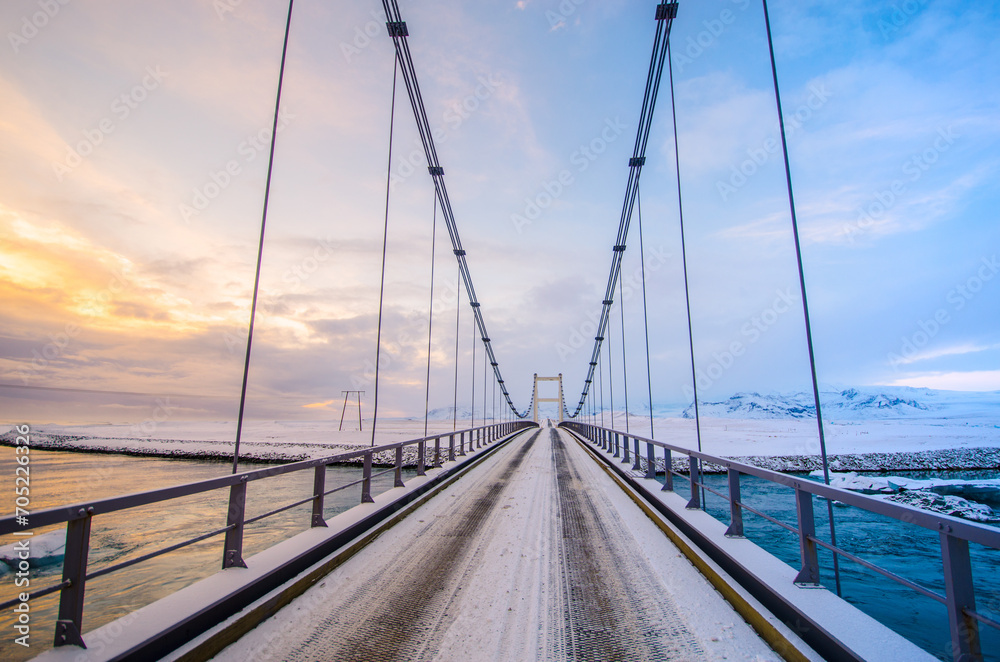 Suspension bridge with steel cables spanning a frozen river against a backdrop of snow-covered mountains at dusk Suspension bridge with steel cables spanning a frozen river against a backdrop of snow-covered mountains at dusk