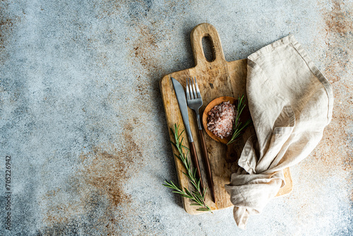 From above rustic kitchen setup on wooden cutting board with a fork, knife, a sprinkle of pink Himalayan salt, fresh rosemary sprigs, and a linen napkin, laid out on a textured concrete background photo
