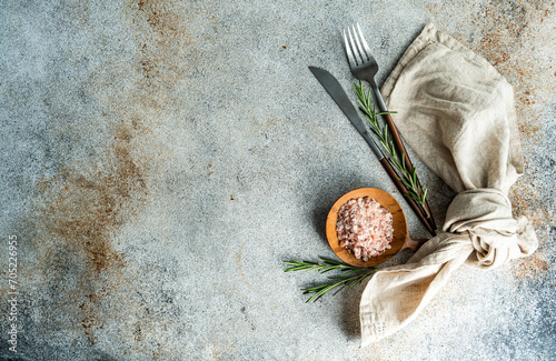 From above wooden spoon with pink Himalayan salt, surrounded by rosemary sprigs and a folded linen napkin alongside a knife and fork, all arranged neatly on a textured concrete surface photo