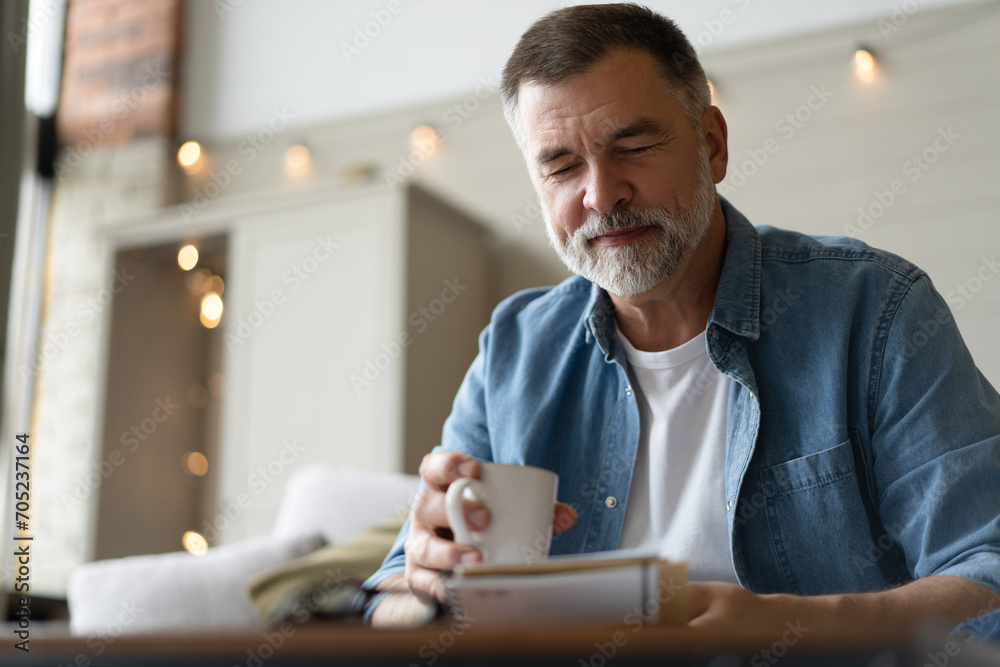 Senior man taking notes in notebook while sitting on the couch, gray-haired elderly man writing thoughts in notebook