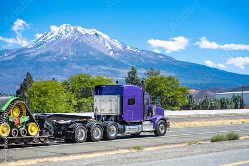 Bright purple powerful classic big rig semi truck tractor with three rear axles transporting equipment on step deck semi trailer running on the highway road with snow mountain on the side