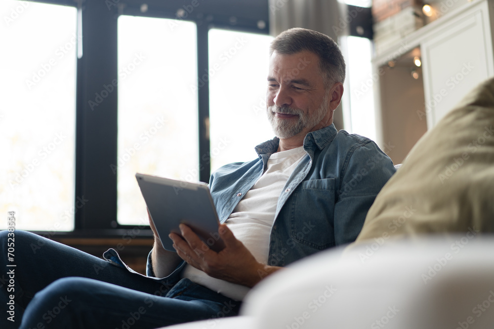Cheerful senior man in casual clothing using digital tablet while sitting on the sofa at home. Cheerful senior man in casual clothing using digital tablet while sitting on the sofa at home.
