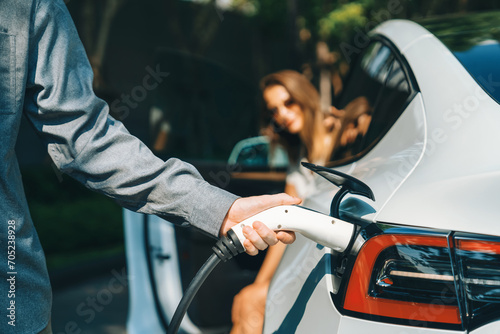 Young couple travel with EV electric car charging in green sustainable city outdoor garden in summer shows urban sustainability lifestyle by green clean rechargeable energy of electric vehicle innards