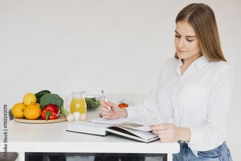 Cute blonde woman is writing something down and looking at a recipe book in the kitchen Cute blonde woman is writing something down and looking at a recipe book in the kitchen