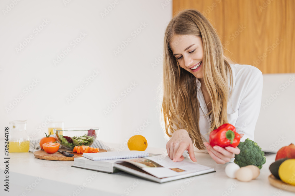Cute blonde woman holding bell pepper and reading recipe book in kitchen on table among cooking ingredients. Cute blonde woman holding bell pepper and reading recipe book in kitchen on table among cooking ingredients.