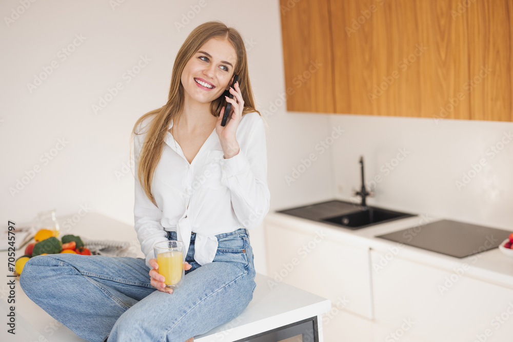 Happy and smiling blonde woman sitting on top of kitchen table drinking orange juice and talking on the phone Happy and smiling blonde woman sitting on top of kitchen table drinking orange juice and talking on the phone