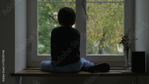 Back of Child sitting by window during winter season looking at snow fall. Melancholic concept young boy daydreaming staring at view from apartment window